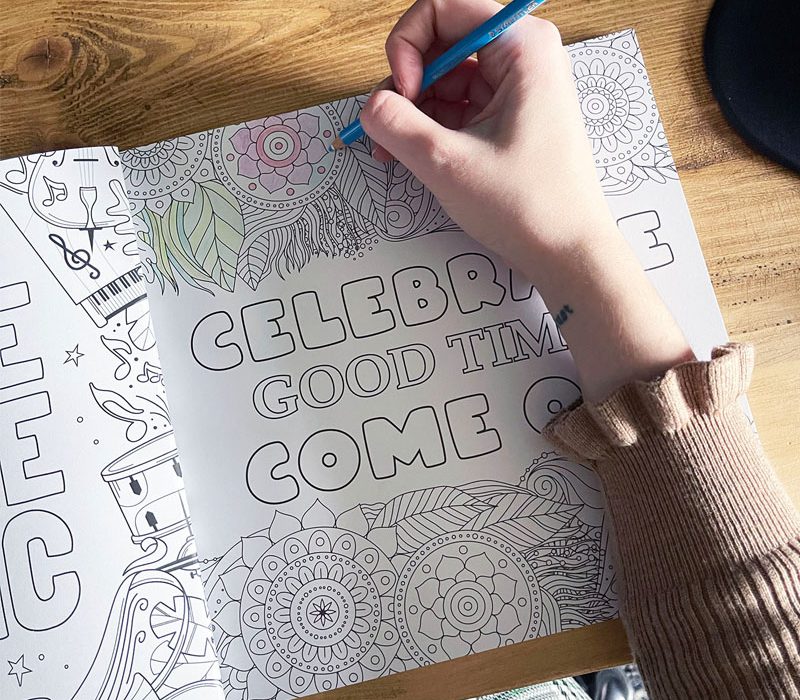 hand holding a blue pencil colouring in an adult colouring book which reads Celebrate The Good Times