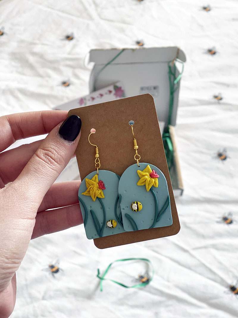 pair of blue arched earrings with daffodils and bees on them. Attached to brown backing card.