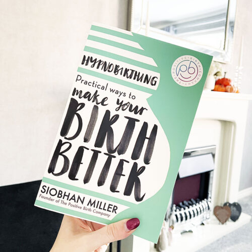 Hand holding a book which reads Hypnobirthing: Practical Ways to Make Your Birth Better, in front of a fireplace.